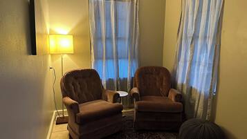 Um quarto com duas poltronas marrons, uma mesa redonda pequena, um tapete estampado e uma luminária.