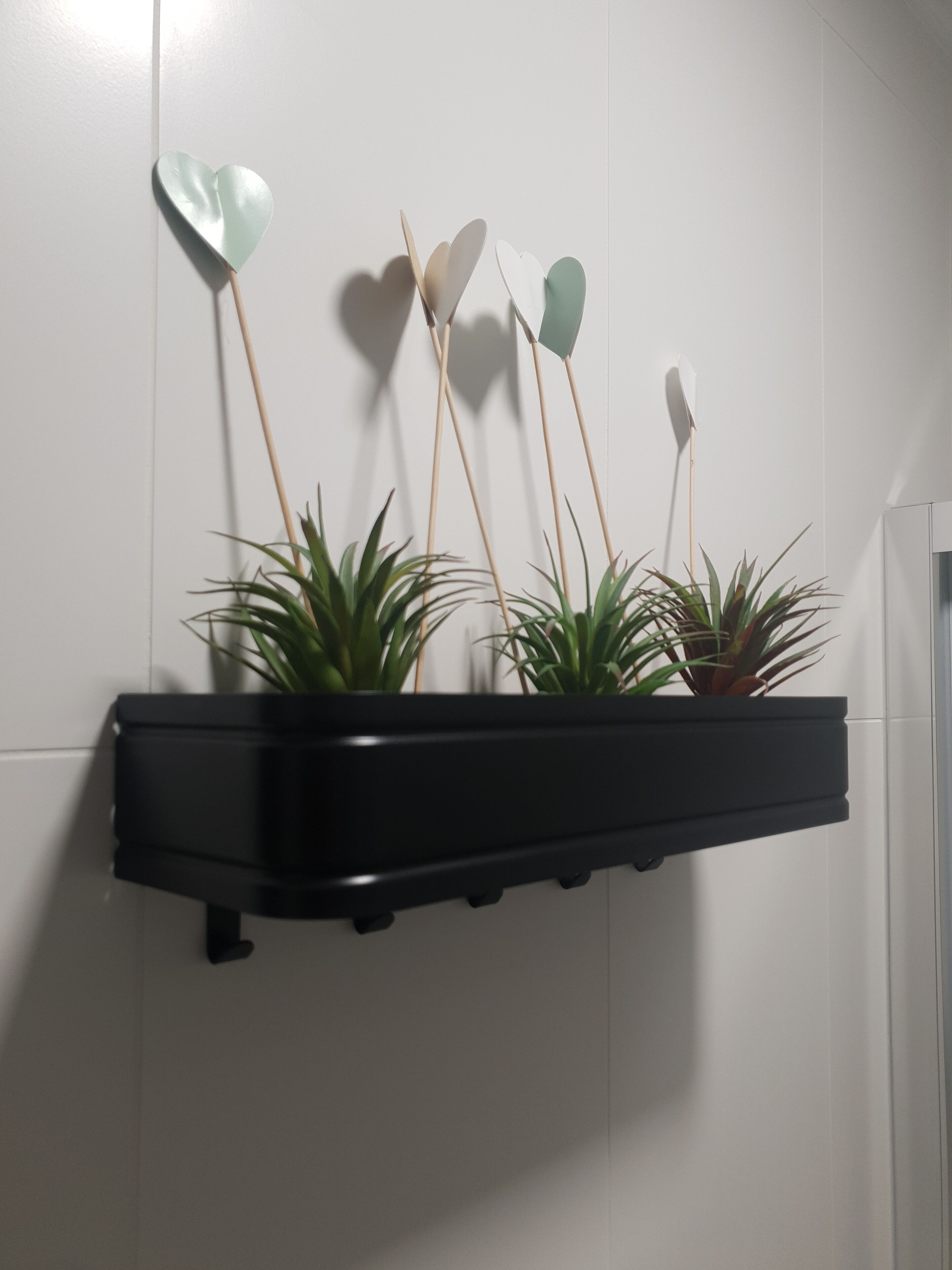 Una estantería negra con tres macetas y adornos con forma de corazón sobre un fondo de pared blanca.