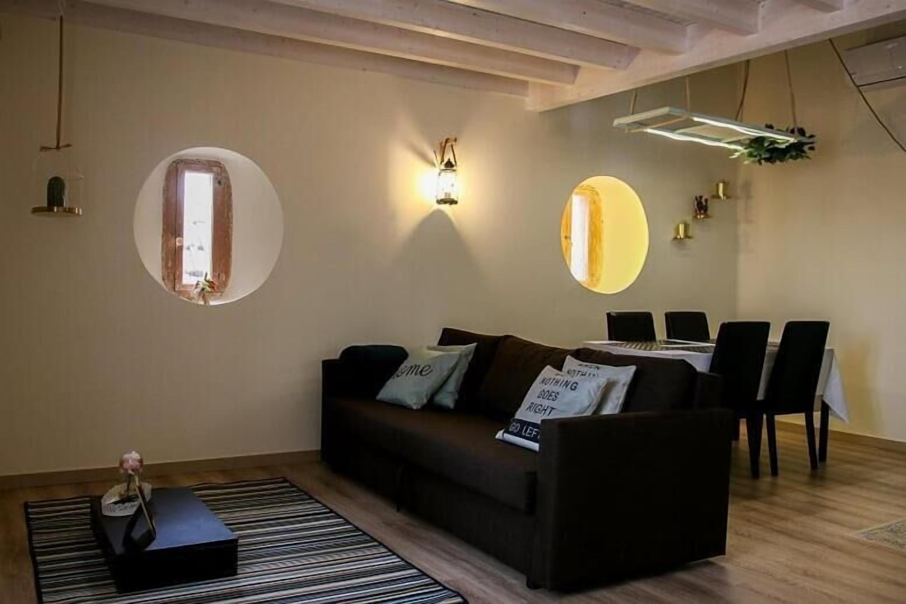 Una sala de estar moderna con un sofá, una mesa de comedor y sillas. Hay un espejo redondo en la pared, un ventanal pequeño y una lámpara colgante.