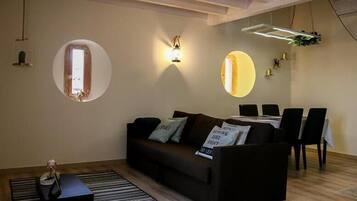 Una sala de estar moderna con un sofá, una mesa de comedor y sillas. Hay un espejo redondo en la pared, un ventanal pequeño y una lámpara colgante.