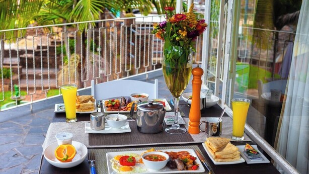 A breakfast table set with various dishes, drinks, and a vase of flowers on a balcony with a view. - Room 1 of 8 Cozy Suites The Hilltop Guesthouse (Harare)