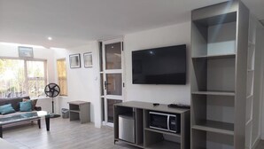 A modern living room with a sofa, a coffee table, a TV, a fan, and built-in shelving. - Room 1 of 8 Cozy Suites The Hilltop Guesthouse (Harare)