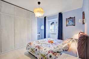 Ein Schlafzimmer mit Bett, Schrank, Pendelleuchte, einem Fenster mit Vorhängen und einem Wandbild.