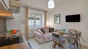 A compact kitchen with a stove, sink, and window. There is a dining table with chairs and a television mounted on the wall. - Ornavasso Apartments (Ornavasso)