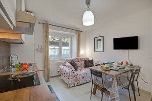 A compact kitchen with a stove, sink, and window. There is a dining table with chairs and a television mounted on the wall. - Ornavasso Apartments (Ornavasso)