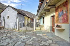 A stone-paved courtyard with a metal gate, a small white chair, and a building with a balcony and a religious painting. - Ornavasso Apartments (Ornavasso)