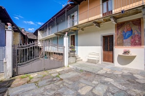 A courtyard with stone flooring, a gated entrance, and a building with a balcony and a door with a religious painting. - Ornavasso Apartments (Ornavasso)