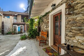Μια αυλή με έναν τοίχο από πέτρα, ένα ξύλινο παγκάκι, ένα τραπέζι με ποτήρια κρασιού και μια πόρτα με τμήμα από γυαλί.