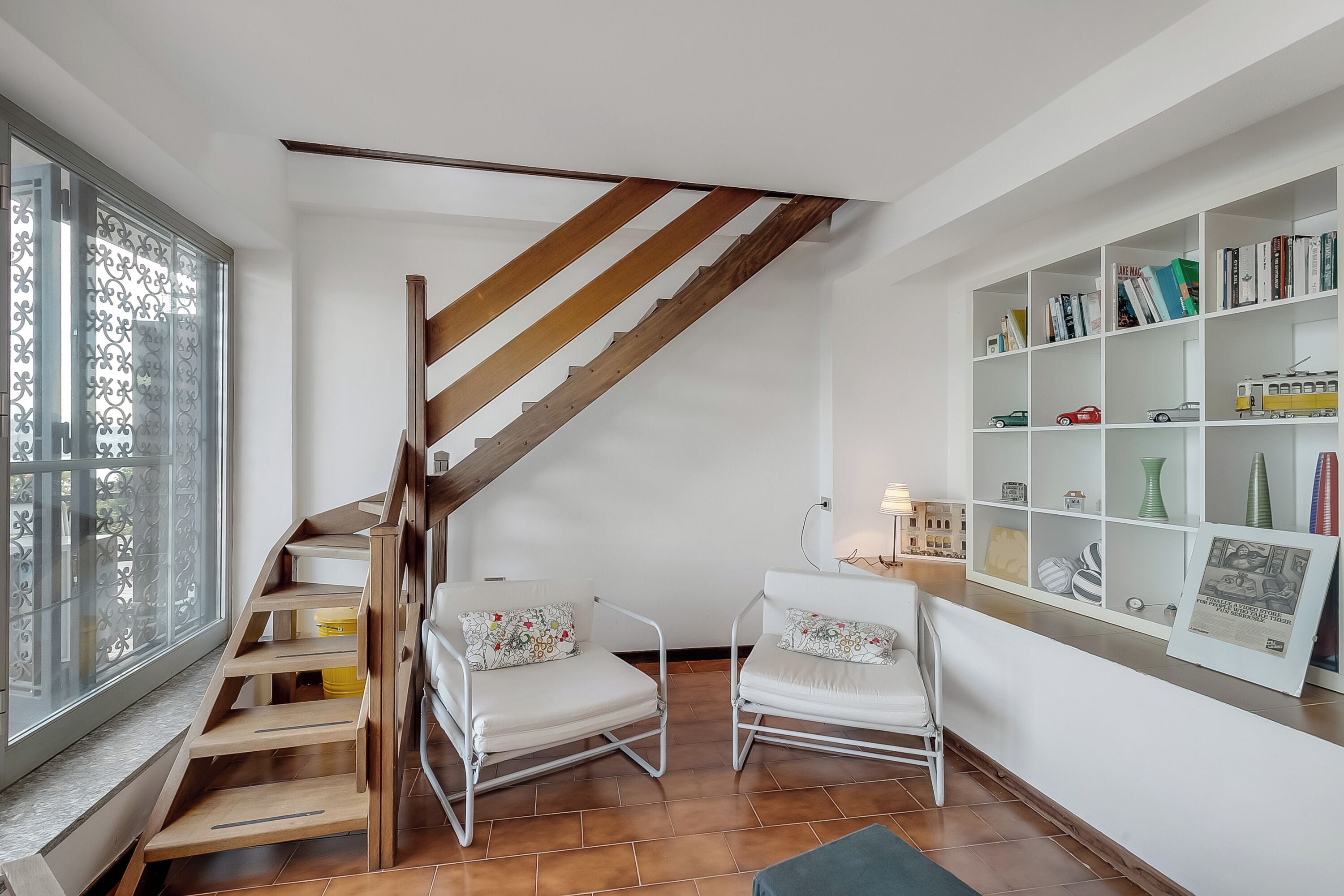 Eine Treppe mit Holzboden und Metallgeländern, zwei weiße Polsterstühle mit floralen Kissen und ein weißes Bücherregal mit verschiedenen Gegenständen.