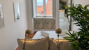 Une chambre avec une grande fenêtre, un lit recouvert de draps blancs, une table de chevet avec une lampe et une plante en pot.