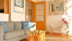 A living room with a sofa, a glass coffee table, a wooden door, a framed mirror, and a small white chair. - Charming 2 Bedroom Apartment w/ River View (Lisboa)