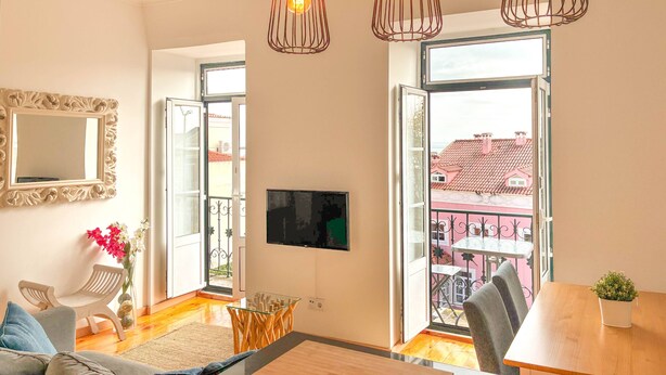 A modern living room with a sofa, a coffee table, a TV, and a balcony with a view of buildings. - Charming 2 Bedroom Apartment w/ River View (Lisboa)