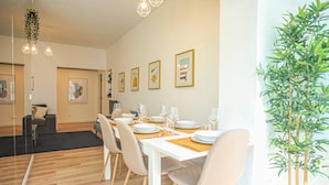 Comedor con una mesa blanca dispuesta para cuatro, rodeada de sillas, con cuadros en la pared y una planta en maceta al lado.