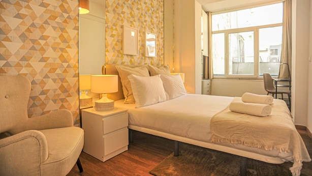 Habitación de hotel con cama, una silla, mesita de noche con lámpara, ventana grande y pared con diseño geométrico.