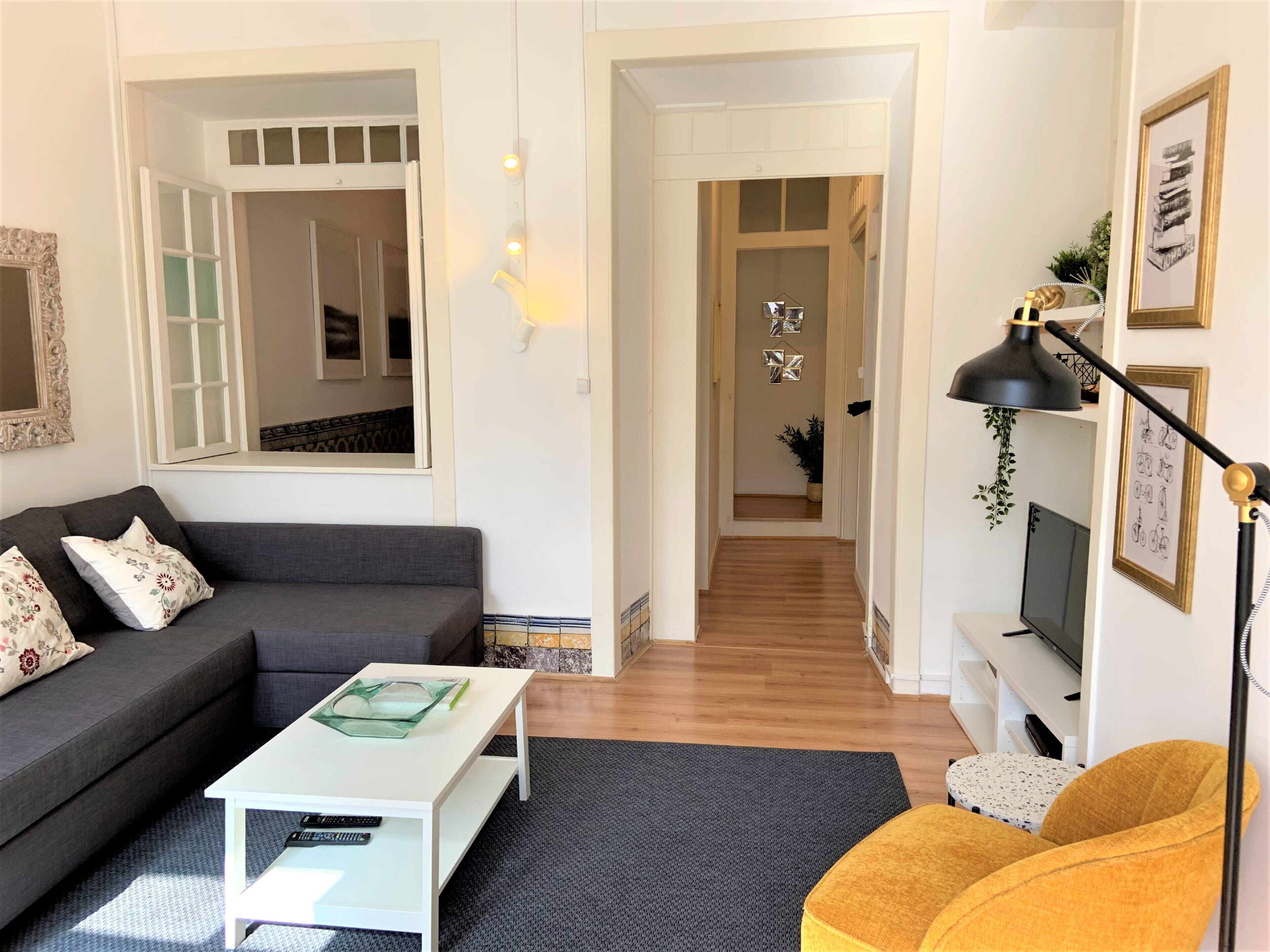 A modern living room with a grey sofa, a white coffee table, and a yellow armchair. There is a TV on a stand and a black floor lamp.