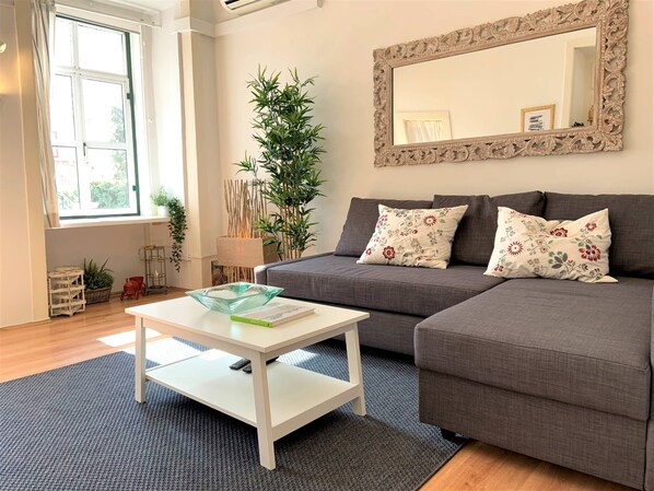 Un salon avec un canapé gris, une table basse blanche, un miroir doté d’un cadre orné et une grande fenêtre.