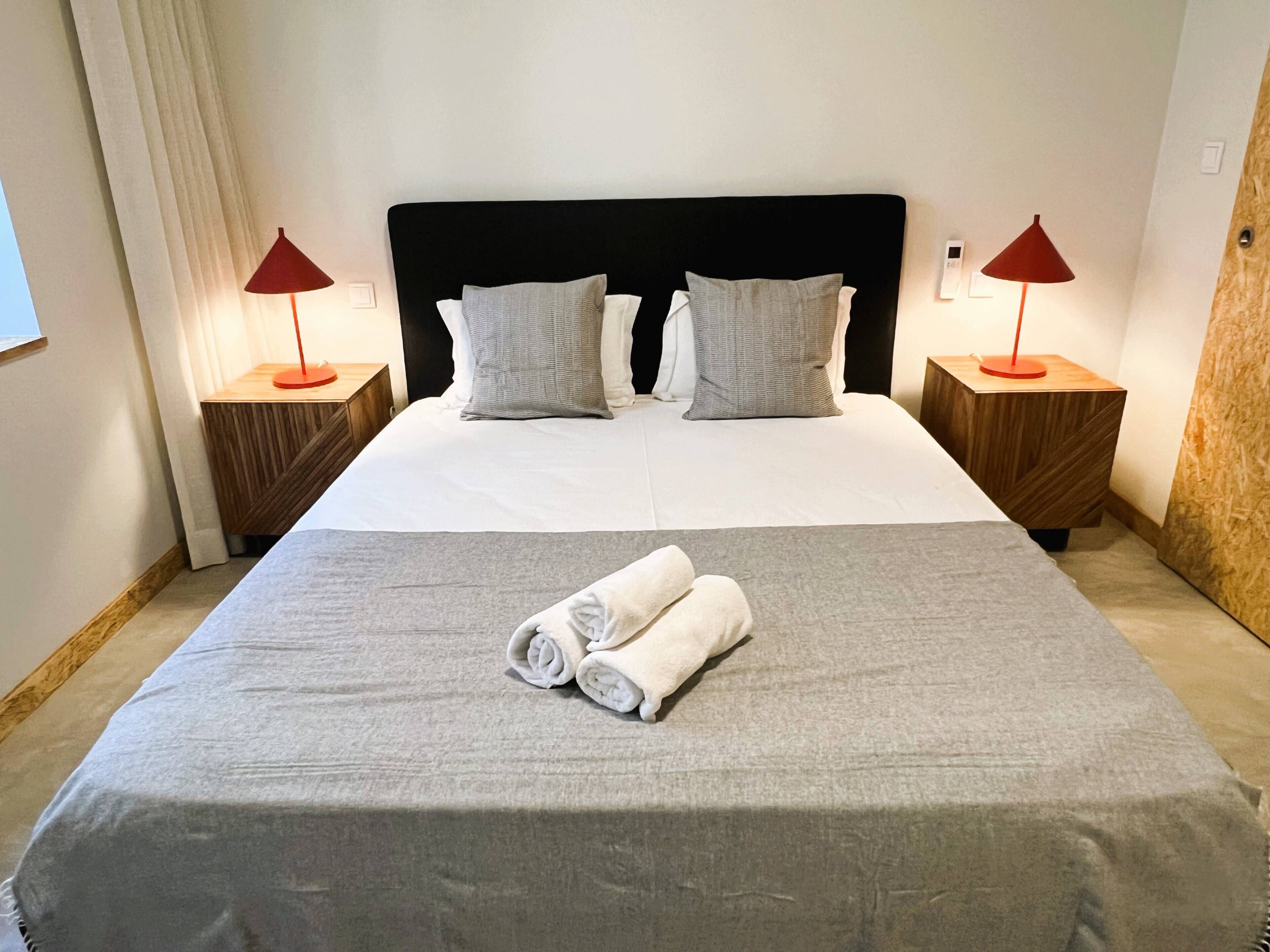 Uma cama bem arrumada com colcha cinza e duas almofadas, duas mesas de cabeceira com abajures vermelhos e uma cabeceira de madeira.