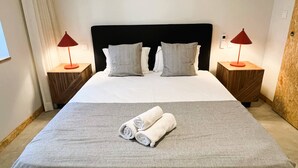 Un lit bien fait, recouvert d’une couvre-lit grise et agrémenté de deux oreillers, deux tables de chevet avec des lampes rouges et une tête de lit en bois.