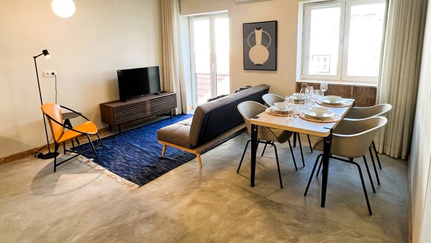 A living room with a sofa, a dining table set for four, a TV on a stand, and a large window with curtains.