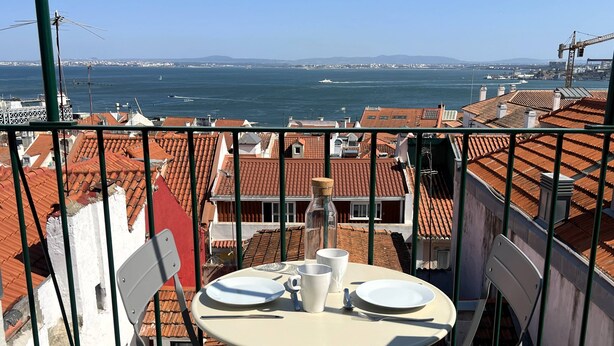 A balcony with a table set for two, overlooking a coastal cityscape with red-roofed buildings and a view of the sea. - NEW Top Modern Loft w/ AC & River View (Lisboa)