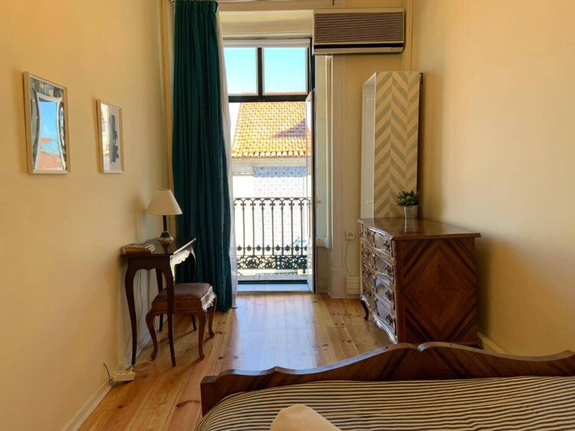 Una camera da letto con un letto, una cassettiera, una scrivania con una lampada e una sedia. Include un balcone con ringhiera e vista su un edificio con tetto in tegole.