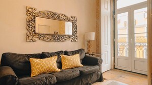 Ein Wohnzimmer mit einem dunkelbraunen Ledersofa, zwei gemusterten Kissen, einem großen, verzierten Spiegel, einem kleinen Tisch mit Lampe und Blick auf einen Balkon durch eine Glastür.