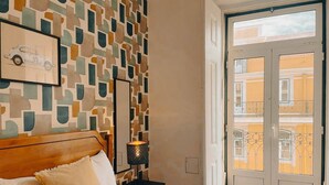 Ein Hotelzimmer mit einem Bett, einem gerahmten Bild an der Wand, einem Nachttisch mit Lampe und Blick auf den Balkon.