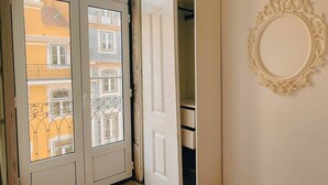 Ein Zimmer mit einer weißen Tür, die auf einen Balkon führt, einer weißen Wand mit einem dekorativen Spiegel und Blick auf benachbarte Gebäude.