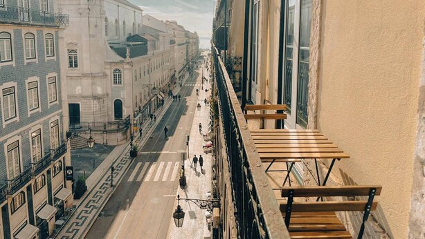 Blick von einem Balkon auf eine Straße mit einer hölzernen Bank, Gebäuden mit großen Fenstern und einer Fußgängerampel.