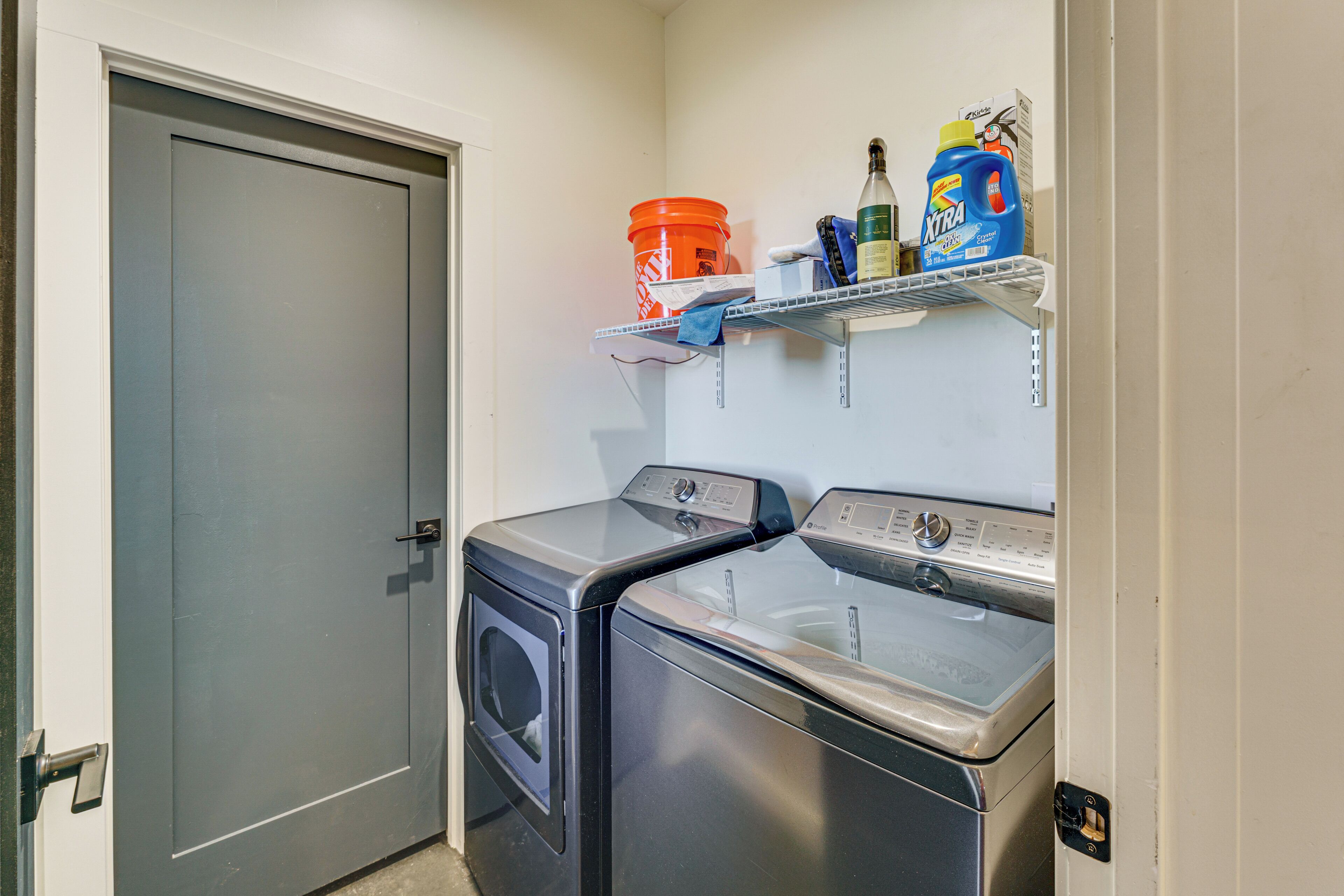 A laundry room with two front-loading washing machines, a shelf with cleaning supplies, and a grey door.