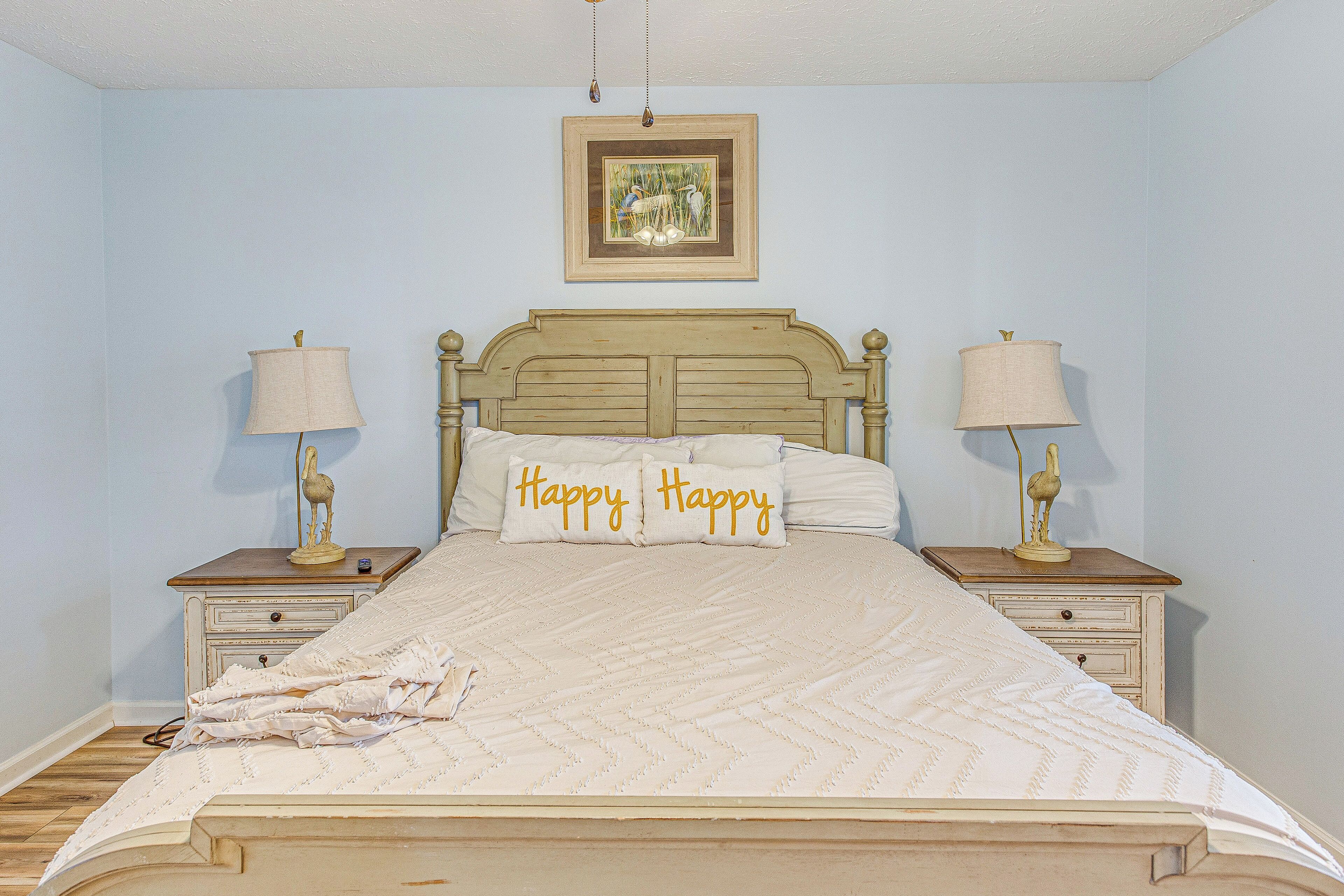 Un dormitorio con una cama, dos mesitas de noche, una lámpara en cada una, un cuadro en la pared y dos almohadas con la palabra 'Happy'.