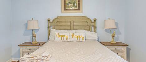 Un dormitorio con una cama, dos mesitas de noche, una lámpara en cada una, un cuadro en la pared y dos almohadas con la palabra 'Happy'.