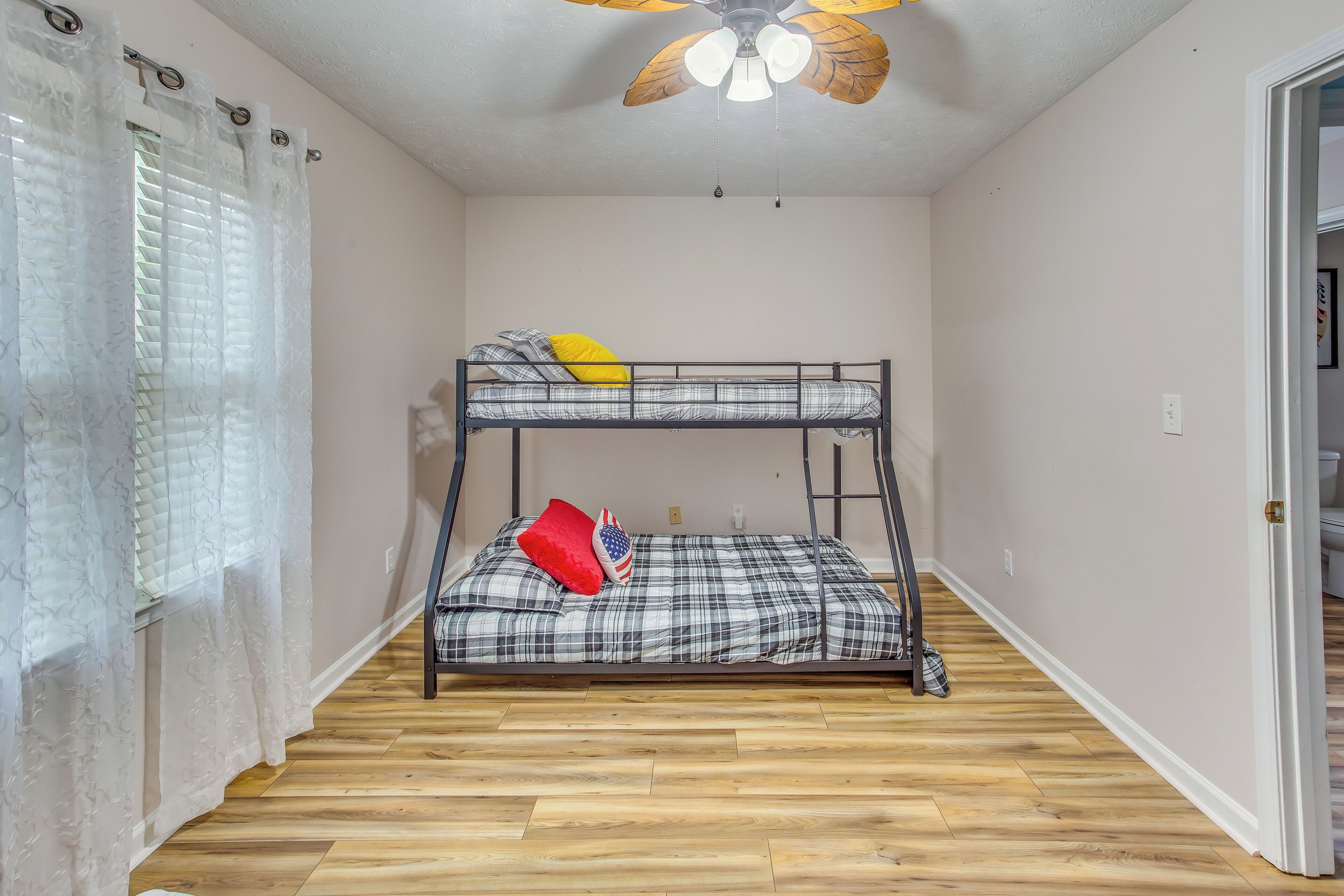 A bunk bed room with a ceiling fan, wooden flooring, and a window with curtains.