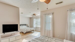 A bedroom with a bed, a TV on a stand, a ceiling fan, and a window with curtains.