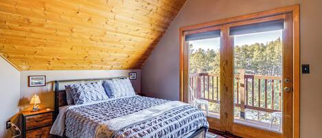 A bedroom with a wooden ceiling, a large bed, a nightstand, a window with a view of trees, and a door to an outdoor deck.