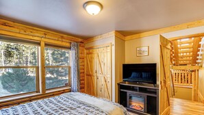 A cozy log cabin bedroom with a bed, a fireplace, a television, and large windows overlooking a forest. - 4 Mi to Fossil Beds: Private Log Cabin on 3 Acres (Florissant)
