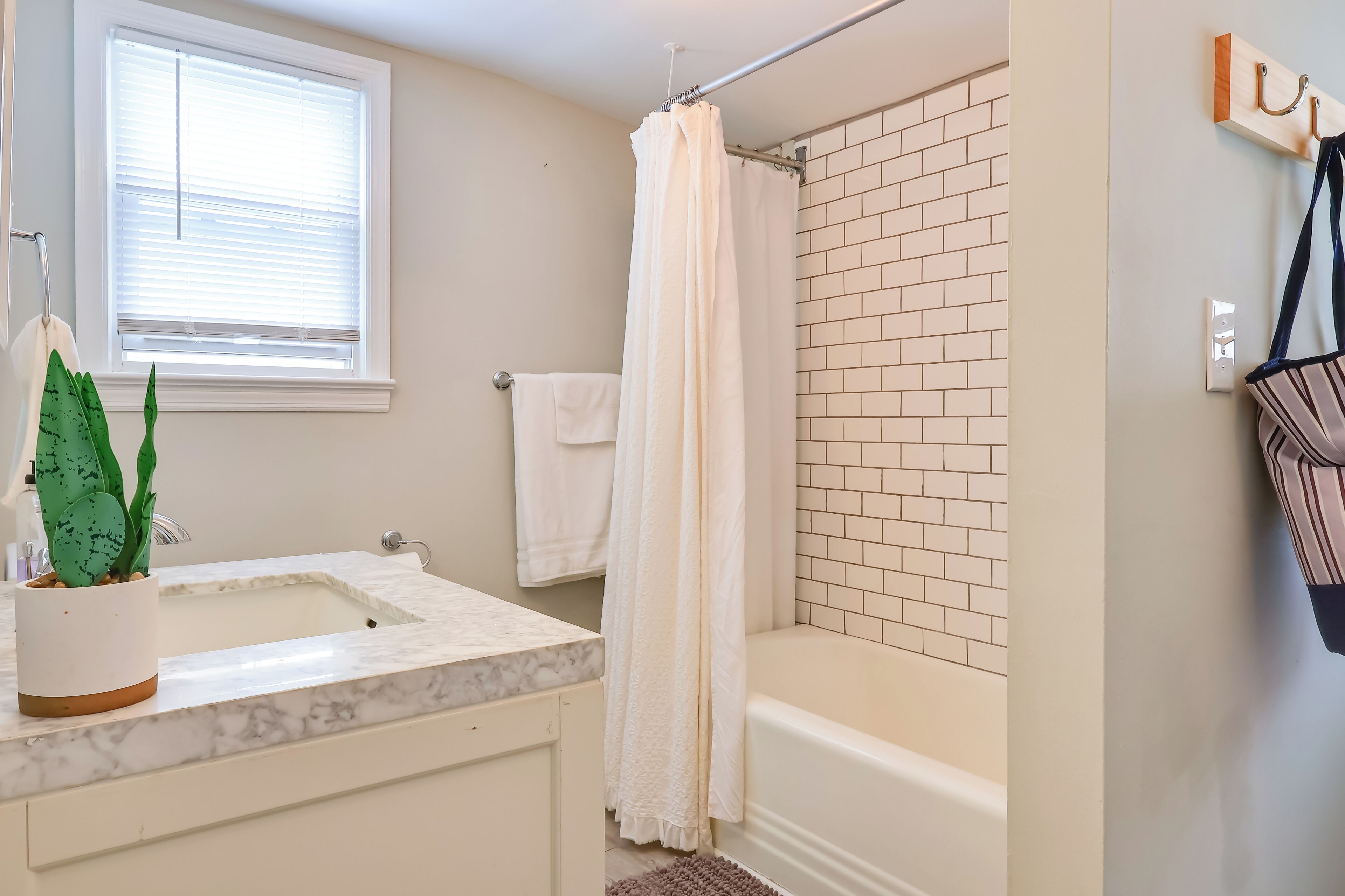 A bathroom with a white bathtub, a white sink with a marble countertop, a potted plant, a window with blinds, and a towel rack.