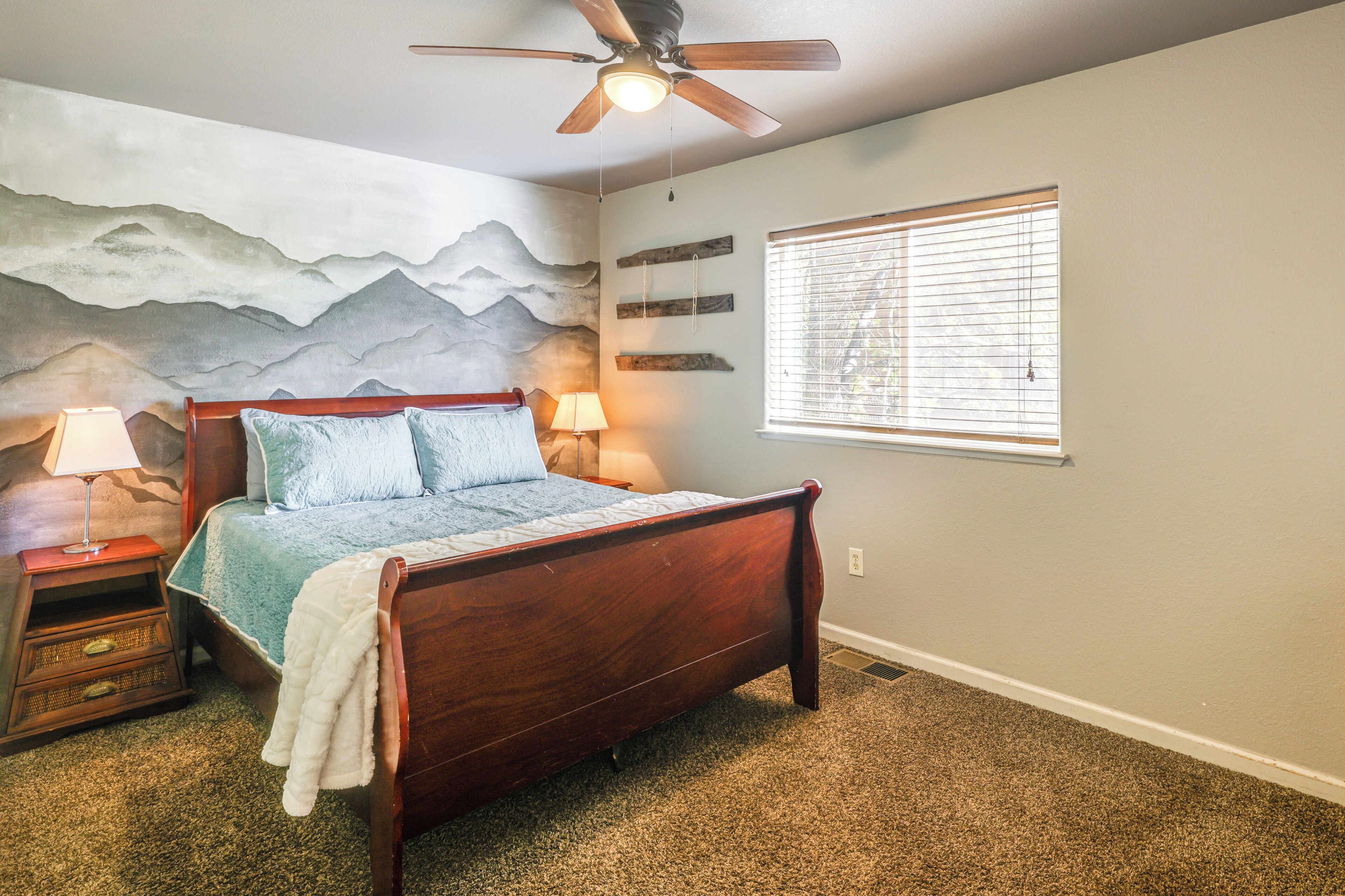 A bedroom with a wooden bed, a ceiling fan, a mountain mural on the wall, and a window with blinds.