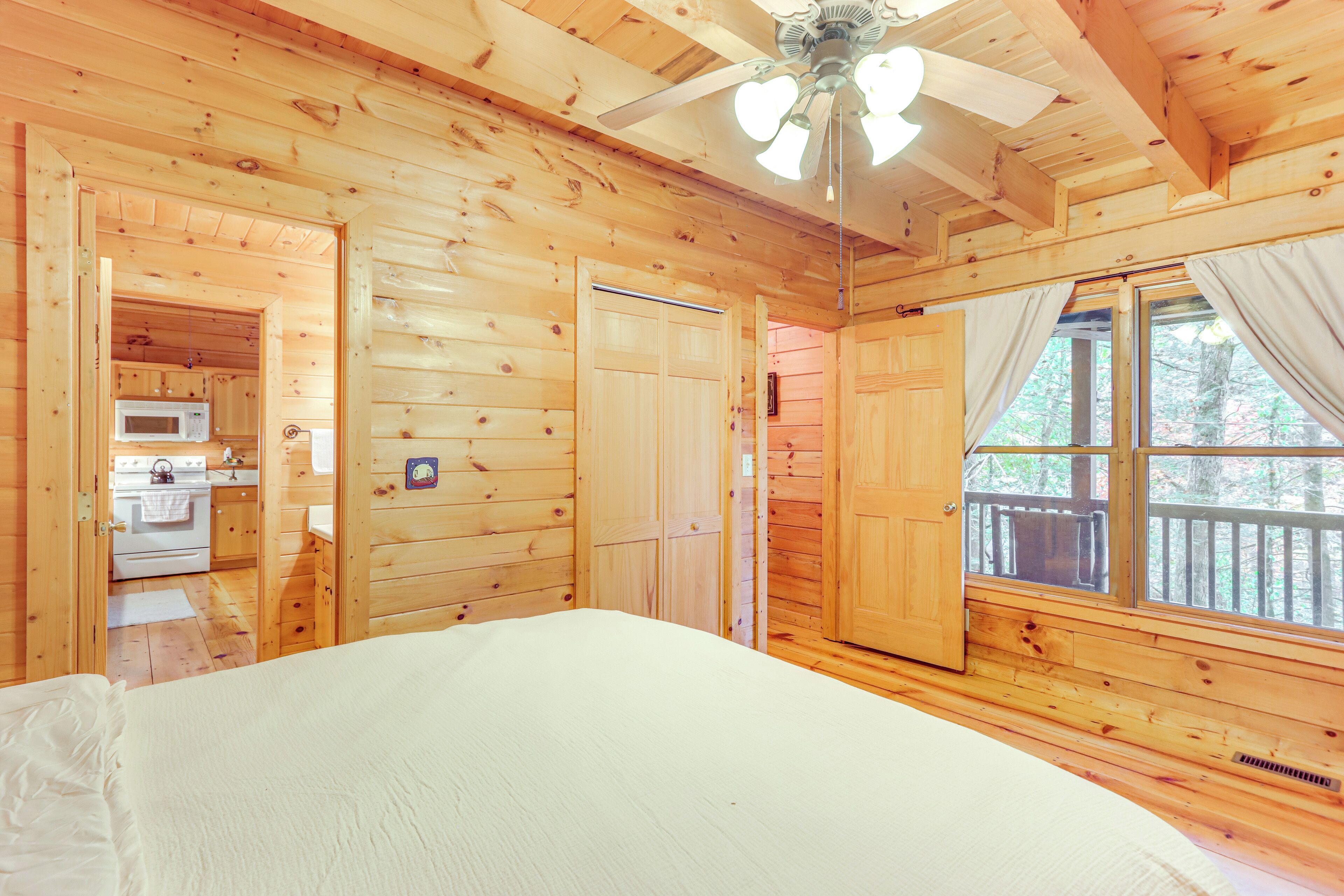 A wooden cabin bedroom with a bed, a ceiling fan, a window with curtains, and a door leading to a kitchen with a microwave.