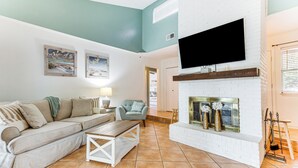 Un salon avec une cheminée en briques blanches, un canapé beige, un fauteuil bleu et un téléviseur à écran plat fixé au-dessus de la cheminée.
