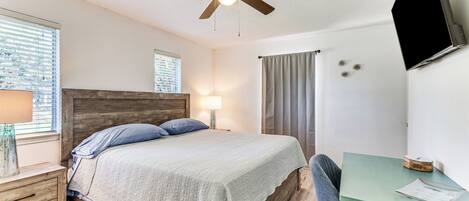 A bedroom with a bed, a desk, a chair, a ceiling fan, a window with blinds, and a TV mounted on the wall.
