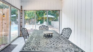 Une terrasse couverte avec une table et des chaises, de grandes fenêtres et une vue sur un jardin avec une piscine et des parasols.