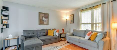 A living room with two grey sofas, a small side table, a floor lamp, and a large window with curtains.
