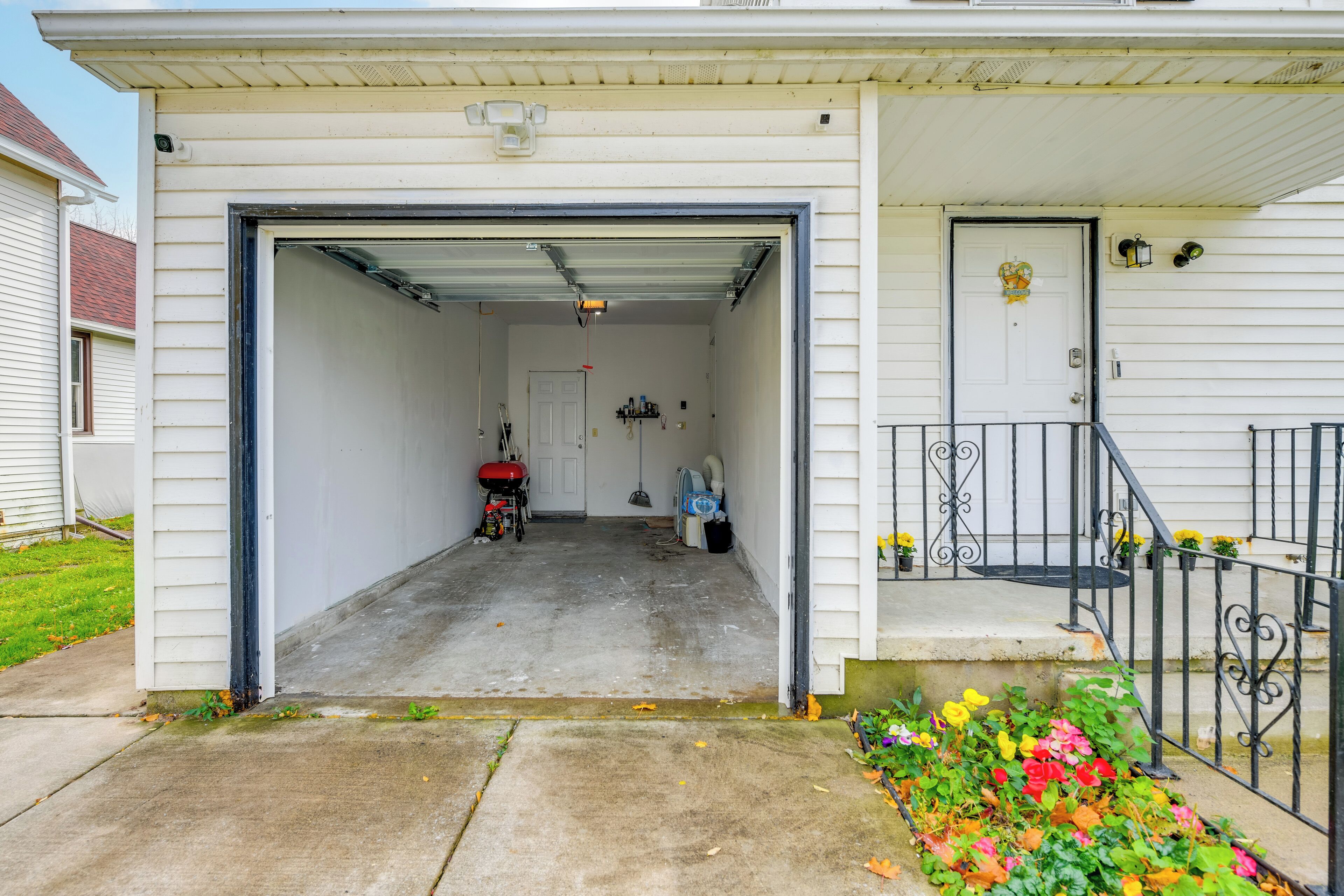 Un garage con un tosaerba rosso e una scopa all'interno, una porta bianca con una ghirlanda e un piccolo aiuola con fiori colorati.
