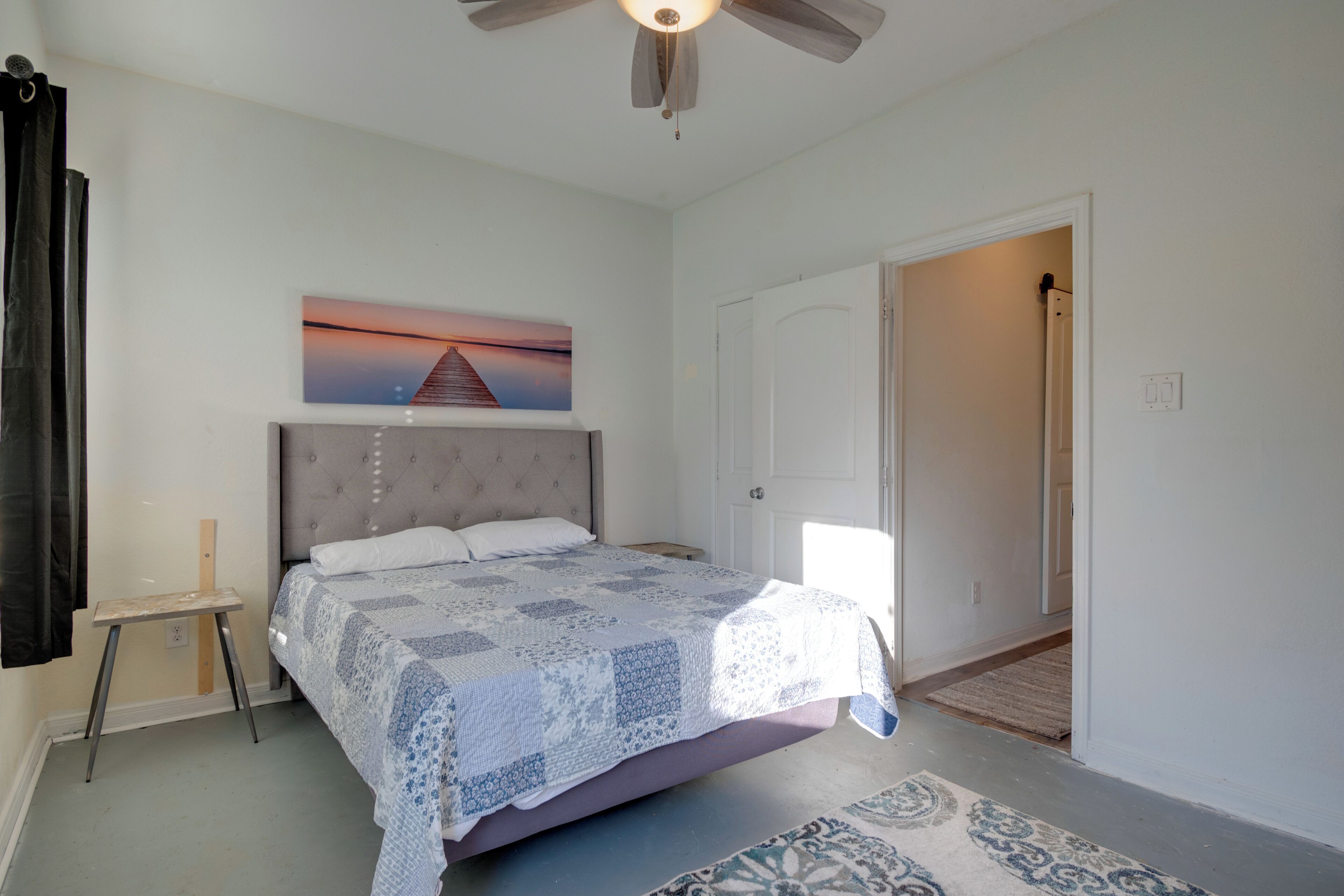 A bedroom with a bed, a ceiling fan, a painting, a door, and a small table.