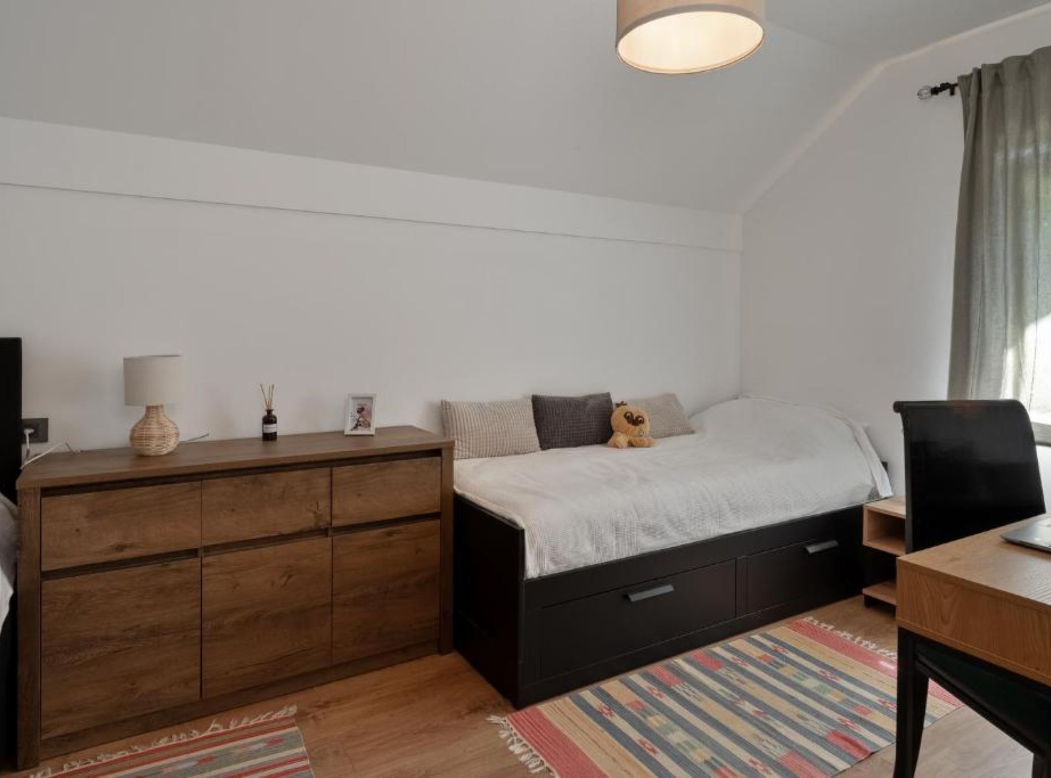 A bedroom with a wooden dresser, a bed with a white comforter, a desk with a chair, and a window with curtains.