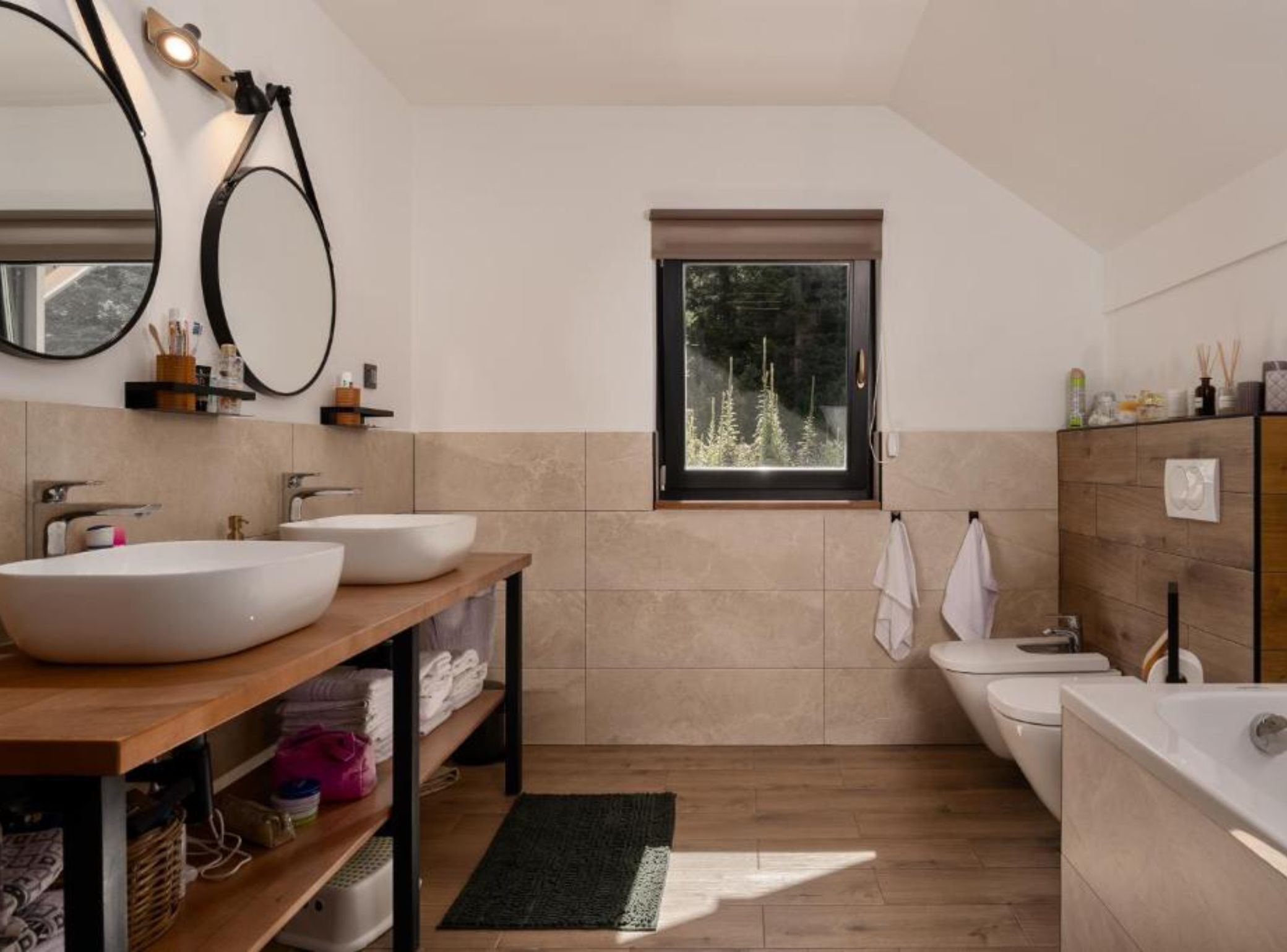 A modern bathroom with two sinks, a bathtub, and a window with a view of trees.