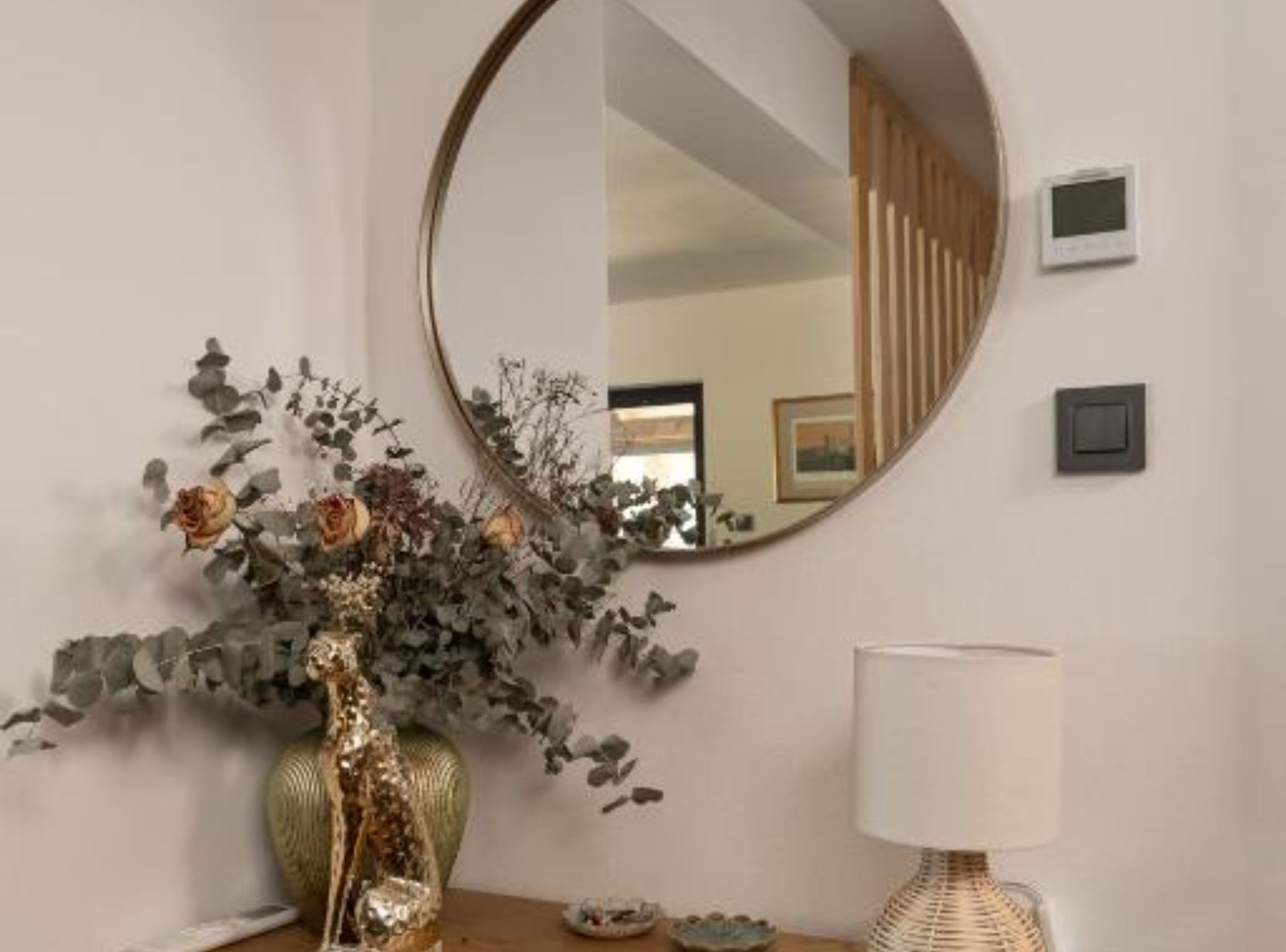 Um quarto com um espelho redondo, um vaso com flores secas, um abajur e uma pequena mesa.