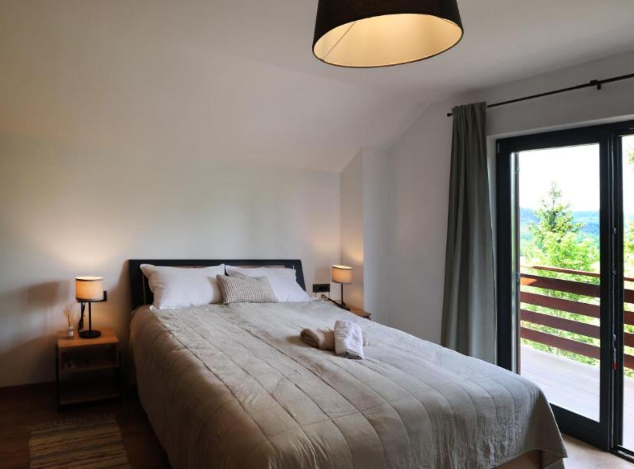 A bedroom with a large bed, two bedside lamps, a view of greenery through a balcony door.