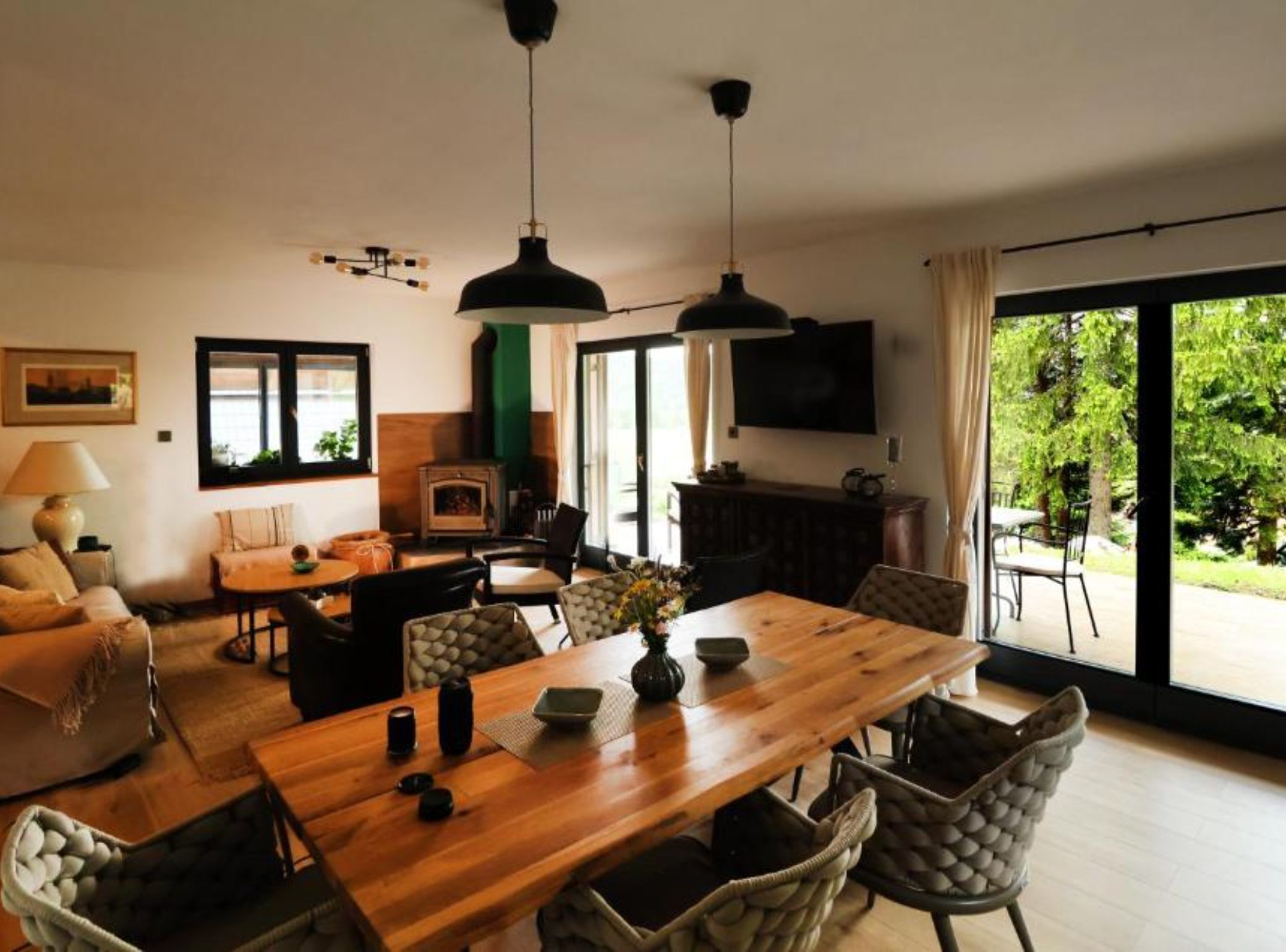 Een ruime woonkamer met een houten eettafel, comfortabele zithoek, een open haard en een groot raam met uitzicht op groen.